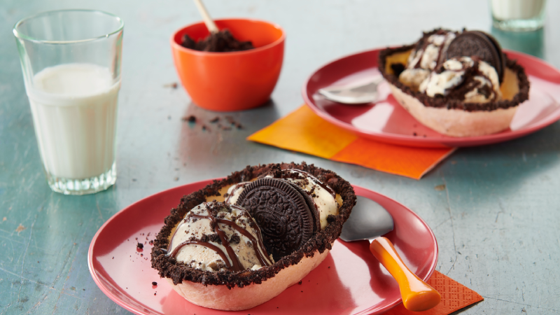 Old El Paso soft tortilla bowls filled with ice cream and Oreos on red plates with spoons, napkins, extra bowl of oreo crumbles and a glass of milk.